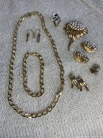 Full set of vintage gold tone jewelry displayed on a light textured fabric background. Includes necklace, bracelet, several pairs of earrings, and a pearl cluster brooch missing its pin.