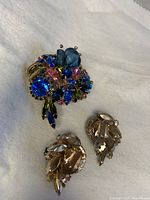 Close-up photo of vintage floral brooch and one clip-on earring showing multicolor rhinestones in blue, pink, and green hues with gold-tone metal base.