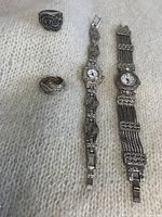 Two vintage silver toned wristwatches and two bold style silver tone rings photographed on a white textured fabric, showing front and band details for the watches and front top views of the rings.
