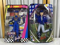 Two Barbie dolls in original window boxes. Left doll in blue NASCAR 50th anniversary race suit holding helmet, right doll in Chicago Cubs baseball uniform and blue jacket with baseball glove. Boxes show aging.
