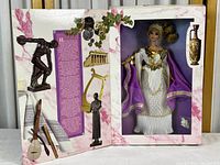 Photo of original box packaging with doll and thematic classical Greek art images and text.