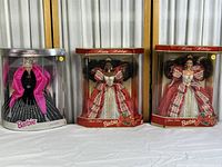 Photo showing three Special Edition Happy Holidays Barbie dolls in their original boxes. Left doll in black and silver gown with pink shawl, middle and right dolls in similar red and white holiday dresses.