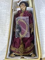 Full view of Sybil, the Flapper doll in original box showing burgundy dress with ornate gold embroidery, dark hair styled in 1920s fashion, and face detail.