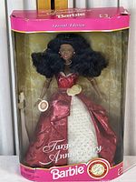 Full front view of boxed Barbie doll showing packaging with pink borders and clear casing. Doll wears red and white gown with gold accents and has curly black hair.