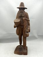 Front view of the hand-carved wooden figurine showing the bearded man with a basket, jug, and walking stick on a base.