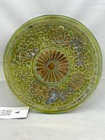 Front view of the 9.5-inch glass plate showing central sunburst motif and surrounding ornate swirling floral scrollwork in amber and olive tones with subtle iridescence.