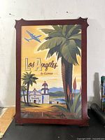 Front view of framed vintage-style Pan Am "Los Angeles by Clipper" travel poster with red beveled frame and decorative cutout corners. Shows tropical trees, Spanish mission building and passenger plane flying.