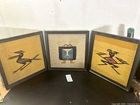 Three framed wool textiles displayed standing on a dark surface, showcasing the geometric folk art figures: a bird, a humanoid face/mask, and a humanoid or zoomorphic figure.