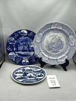 Three blue and white antique transferware plates arranged standing and lying on a gray surface, showing different decorative scenes and scalloped edges, displaying designs and patina
