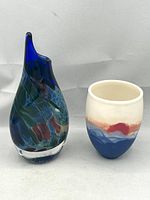 Photo showing full view of a blue teardrop-shaped glass vase with swirling colors next to a small porcelain cup with blue, red, and cream glaze.