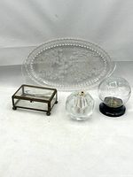 Photo showing four vintage glass and metal decorative collectibles: etched glass oval tray, cut-glass match striker, molded glass baseball coin bank, and beveled glass and brass trinket box grouped together on white background.