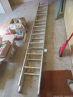 Full view of folded aluminum extension ladder laid on floor near boxes and cleaning broom.