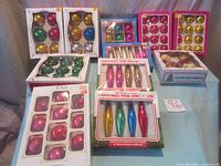 Overall view of 11 boxes containing various glass Christmas tree ornaments in multiple colors and shapes on blue cloth background