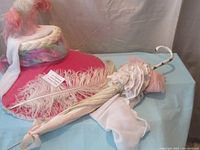 Pink velvet and chiffon bonnet decorated with ostrich feathers lying next to a parasol on a blue cloth background.