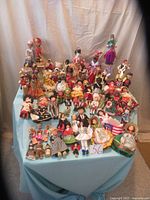 Photo showing a large collection of international dolls arranged on a three-tiered blue cloth-covered display. Dolls represent many countries, wearing cultural outfits with varying sizes and colors.