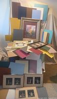 Overview photo showing several sheets of matboard in multiple colors and sizes stacked and leaning against a surface, along with multiple framed pictures including an inspirational picture and cross-stitch lighthouse pictures.