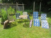 Overview showing two blue-and-white woven strap lounger chairs, two folding chairs with green and white strap seats, small round metal table, folded patio umbrella, and stack of eight beige folding chairs with lattice pattern.