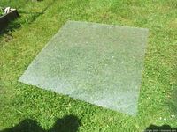 Single large clear plexiglass sheet laid on green grass, square shape, minimal warping, moderate surface marks.
