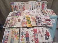 Wide view showing about 35 sewing patterns arranged on a flat surface. Various brands and types depicted with illustrated garment designs on covers.
