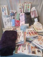 Collection of sewing patterns displayed standing and laying flat, with various fabrics and trims surrounding them. Shows diversity of pattern types and fabric textures/colors.