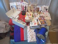 Wide view of sewing patterns, assorted fabrics, Pfaff plotter accessory, accessories storage box, sewing machine bobbins, and other sewing items displayed on a surface.