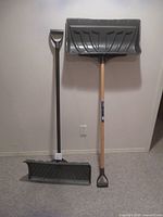Two snow shovels standing upright against a wall, both with wooden handles and grey molded plastic scoops, one with a D-grip handle and the other with a straight handle.