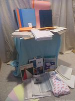 Photo showing assorted duo-tang folders in blue, orange, pink, and black colors on top of piles of white papers and envelopes with some paper packs and plastic sleeves at the front.