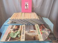 Photo showing 20 hardcover Stitch By Stitch books arranged side by side and three books laid flat in front displaying cover art of homemade dresses and quilts.