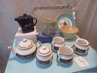 Full lot overview showing dark blue Tetley teapot, cream colored decorated tea set including teapot, milk jugs, sugar basins, handmade ceramic plant pot, Christmas plate, and aluminum tray