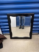 Square vintage mirror with decorative carved edges on the black painted wooden frame against a blue storage door background, showing front reflective surface.