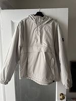 Front view of men's light beige hooded jacket by Pull and Bear, showing hood, elbow length zipper, front pockets, elastic cuffs, and logo patch on left sleeve.