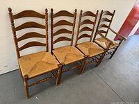 Front angle of four solid wood ladder back chairs with woven seats, arranged in a row, one chair with armrests.