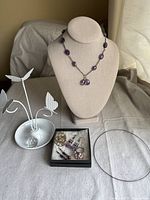 Amethyst necklace displayed on bust stand, three pairs of earrings in black box, pink beaded adjustable ring, and silver metal butterfly earring display on table.