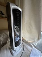 Front view of the Area Max air cleaner tower unit with control panel visible. The unit is white with a black glossy front control panel showing buttons and indicators. It is standing upright on a flat surface near a window with a curtain.