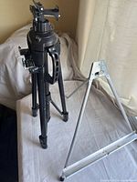 Photo showing black collapsible tripod next to white metal tabletop easel on a white surface with draped fabric background
