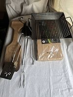 Photo showing the full set of BBQ tools including wooden paddle, metal spatula, forks, grilling basket, and wrapped cedar planks.