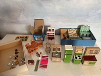 Photo showing full set of 1970s vintage dollhouse miniatures including kitchen and bedroom furniture, dining table set, small accessories on white surface against a painted background.