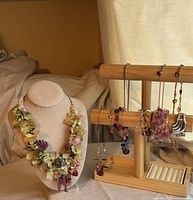 Photo showing the floral statement necklace prominently placed on a bust display with assorted beaded necklaces and bracelets hanging from a wooden jewelry stand in the background.