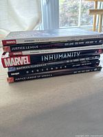 Stacked view of the seven hardcover comic books showing titles and publishers on spines: Avengers Time Runs Out Volumes One and Two, Justice League Injustice League, Marvel Inhumanity, Batman/Superman Second Chance, Image Descender, Justice League of America.