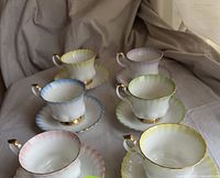 Six Royal Albert bone china tea cups and saucers in pastel colors of yellow, blue, pink, green, and purple, with gold rims, arranged on fabric
