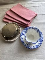 Tea cup with matching blue floral saucer, extra saucer, vintage metal coaster set, and four pink linen napkins arranged on beige fabric.