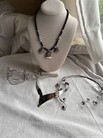 Front view of black beaded silver tone necklace with ornate pendants and three silver tone bracelets on beige cloth with curtain background.