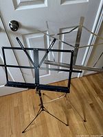 Black metal frame collapsible music stand set up in front of a white door, shown with foldable supporting arms and tripod base.