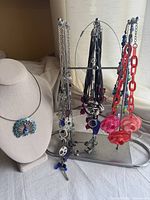 Photo of multiple statement necklaces on a display stand and peacock pendant necklace on bust. Shows variety including leather tassel necklaces, chain necklaces with charms, and large resin link necklaces with flowers.