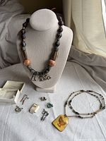 Photo showing handmade heavy beaded necklace with a metal chicken pendant, chain necklace with yellow tin chicken pendant, and various pairs of clip-on earrings arranged on a table.