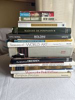 Stack of vintage art books arranged on a white surface, titles visible including Art Treasures of the Louvre, Prado Gallery, Becoming Mona Lisa, Madame de Pompadour, Boldini by Vito Doria, among others.