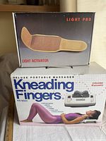 Two vintage body work appliances in original boxes, a Kneading Fingers massager and Light Activator light pad, showing front packaging and scale.