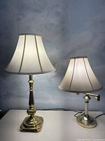 Two brass lamps on a white surface against a muted background. The taller lamp on left with a stepped base and pleated bell shade; shorter lamp on right with round base and swinging arm.