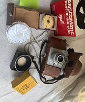Photo showing Kodak Pony 135 camera in brown leather case, Unittic light meter, GAF Transcomatic Developing Tank box, Kodak flash bulbs in yellow box, light diffuser, and camera hood with strap arranged on surface.