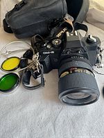Vintage black Chinon CM-5 camera with attached 35-80mm zoom lens, black camera strap with Olympus branding, yellow and green lens filters, and carrying case shown on a fabric surface.